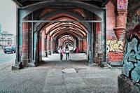 two people walking down a street under an arched bridge